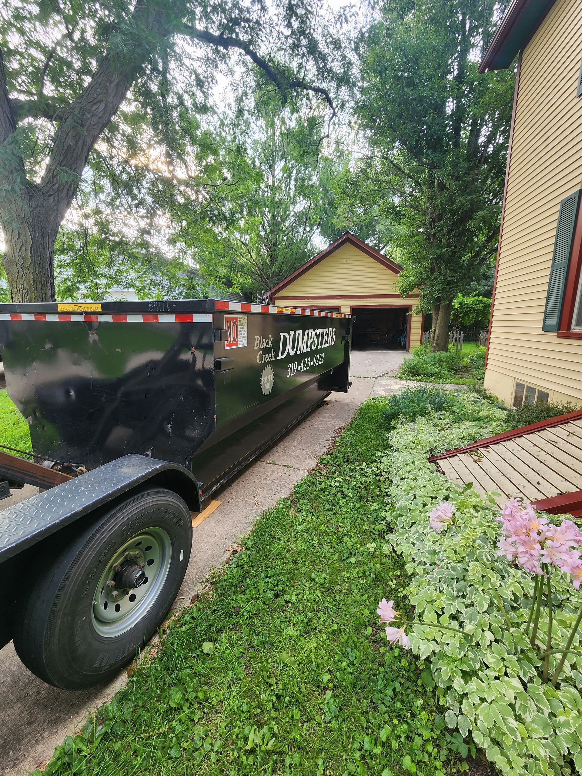 Black dumpster on a trailer parked on a driveway next to a house with green bushes and a garage.