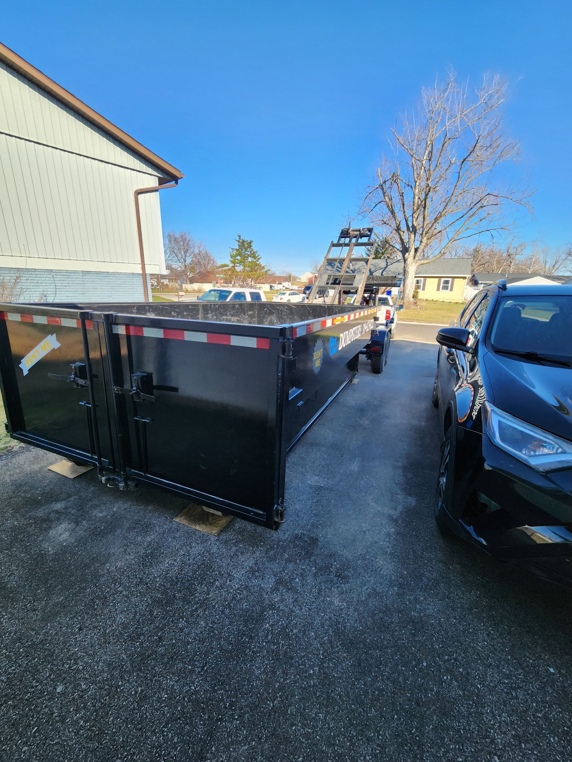 Black dumpster on driveway next to a black car.