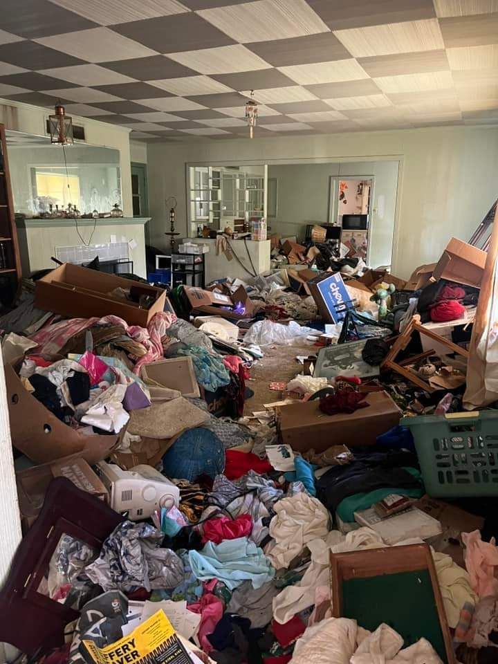 Cluttered living room with boxes and piles of clothes and objects.