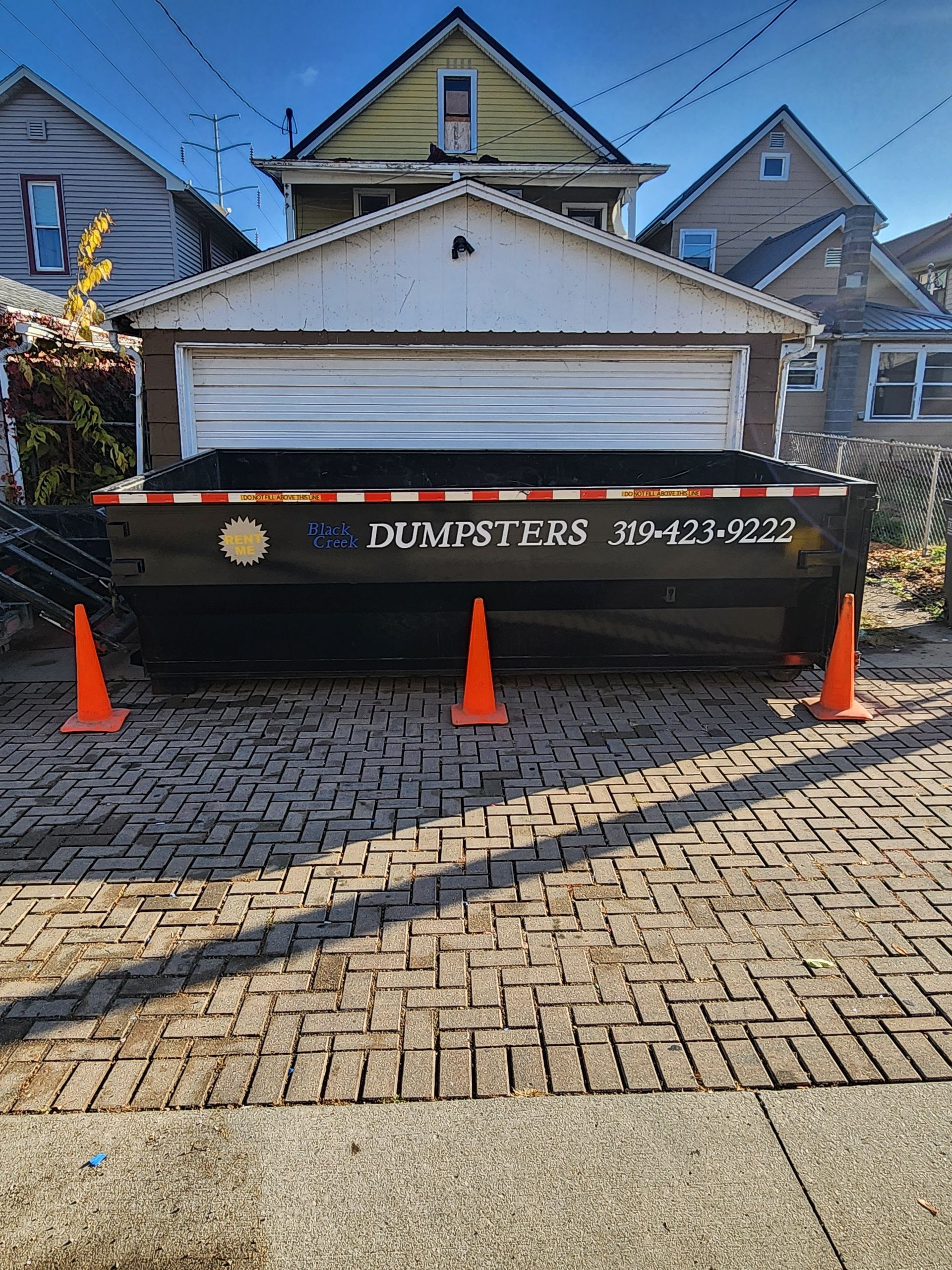 Black dumpster on brick driveway, orange cones, garage in background.
