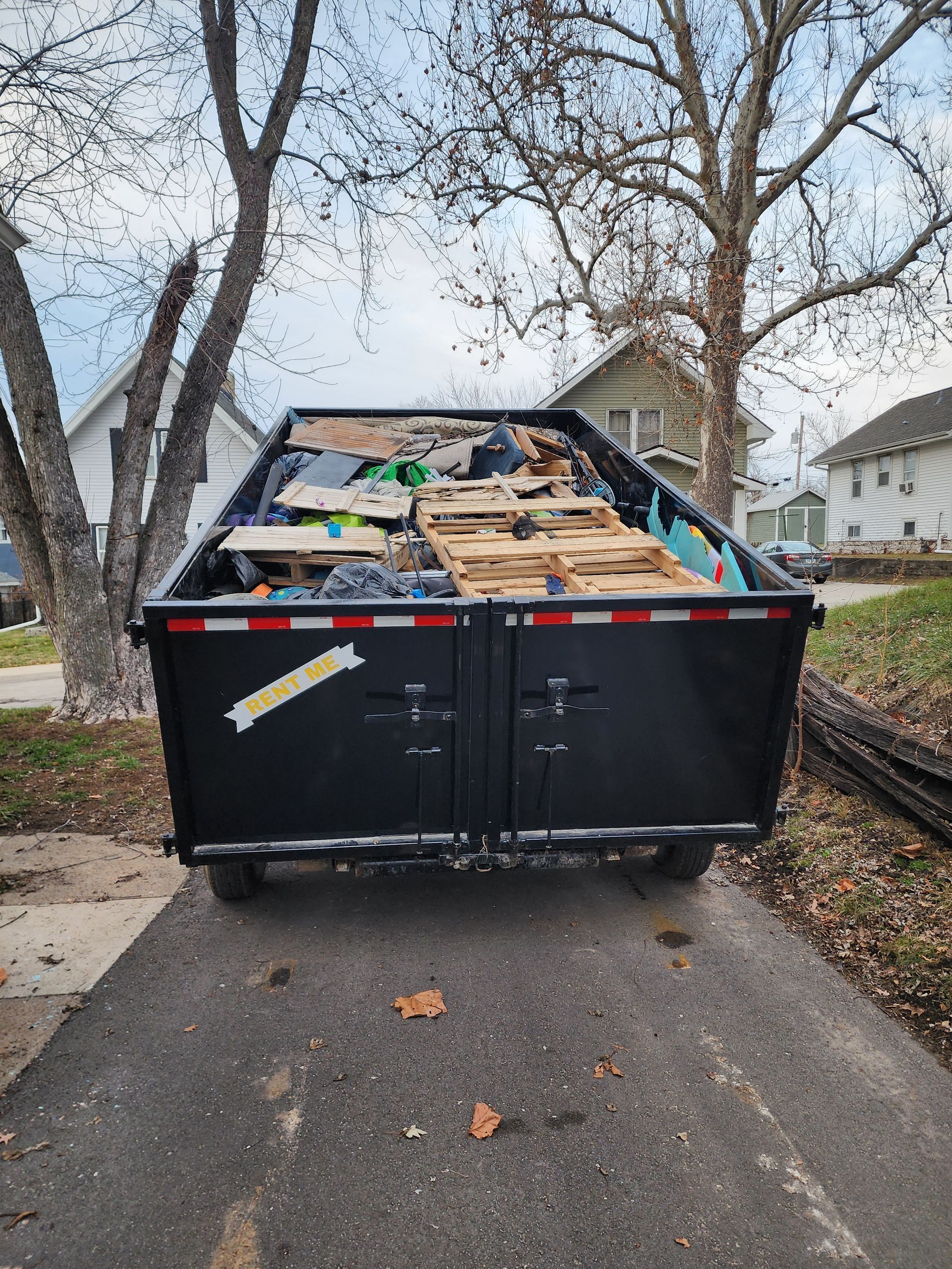 Black dumpster on a driveway filled with trash, in front of houses and trees.