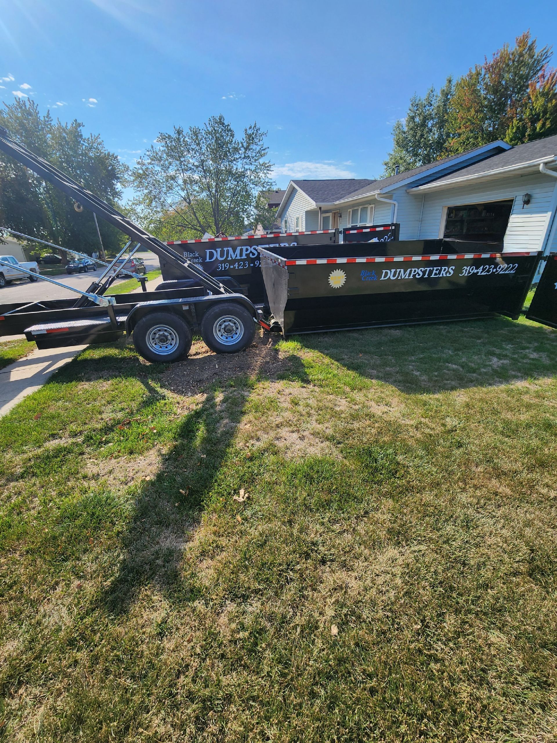 A large black roll-off dumpster trailer parked on grass beside a house.