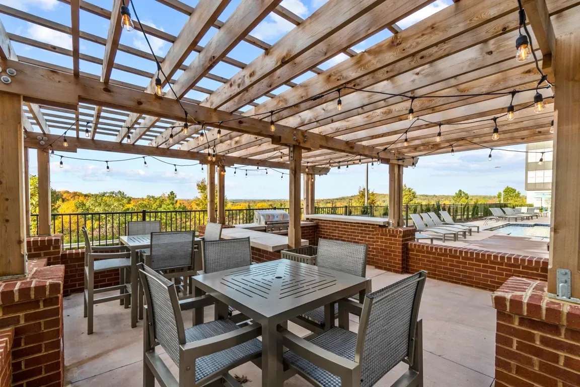 Outdoor seating area with tables and chairs under a wooden pergola with string lights, overlooking a pool and lounge chairs.