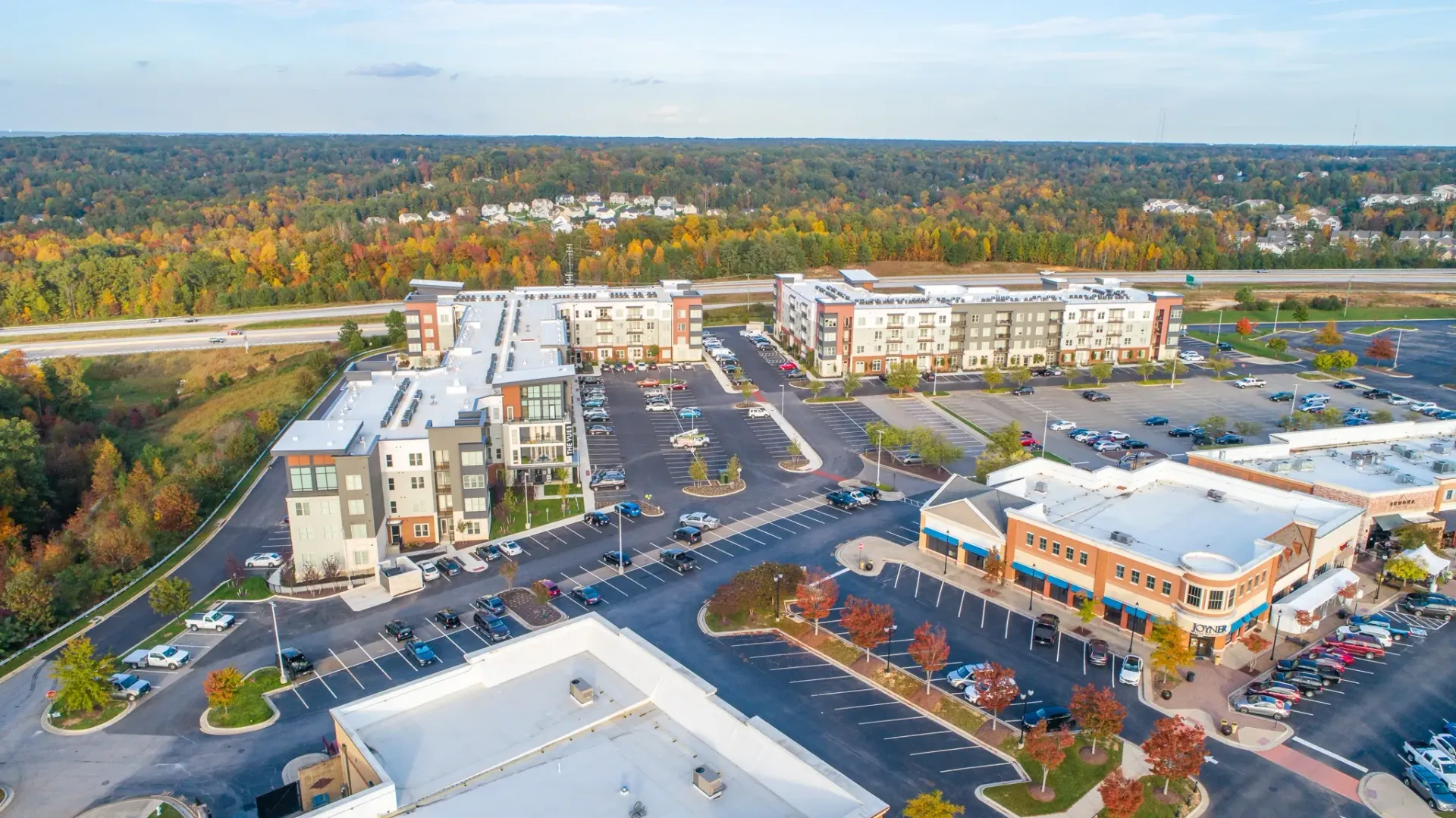 Aerial view of apartment buildings and a retail strip mall with a highway and autumn trees in the background.