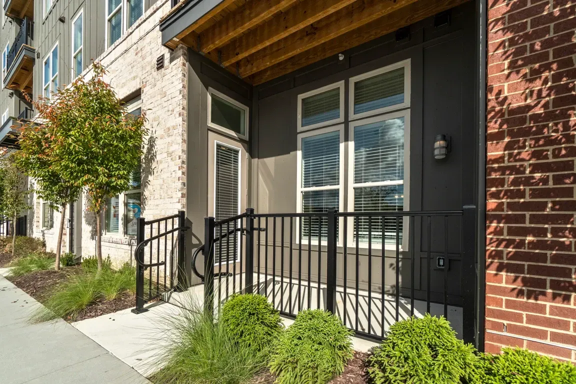 Entrance to a modern apartment building with brick and dark siding, featuring a private balcony or patio area with a railing.