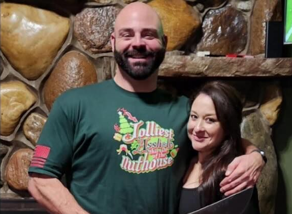 Man with a beard and woman pose in front of a stone wall. Man wears a green shirt. They are smiling.
