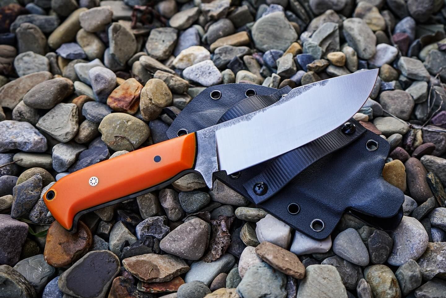 Orange-handled survival knife with a serrated blade in a black sheath, resting on a bed of rocks.
