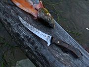 Fish fillet knife next to a fish head on a weathered wooden plank near water.