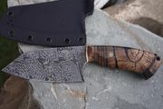 A handmade cleaver knife with a patterned blade and wooden handle, resting on a gray rock with a black sheath.