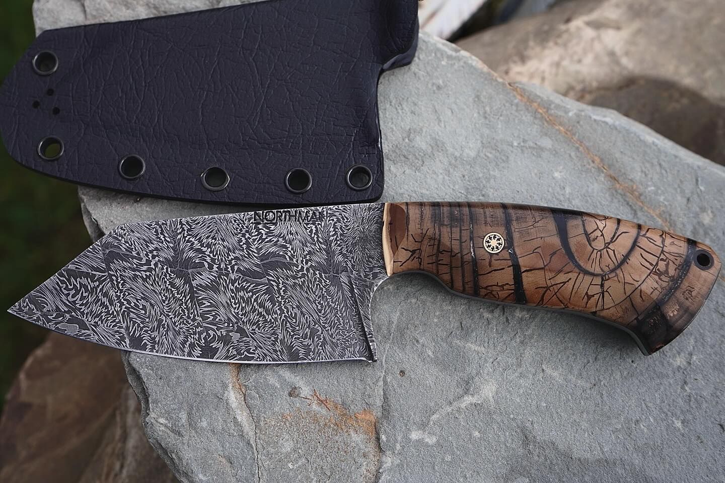 Damascus steel cleaver knife with wood handle and black sheath on a rock.