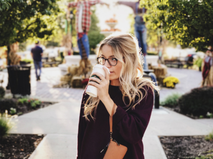 Blonde woman wearing glasses sipping from a coffee cup outdoors; fall scene with decorations and trees.