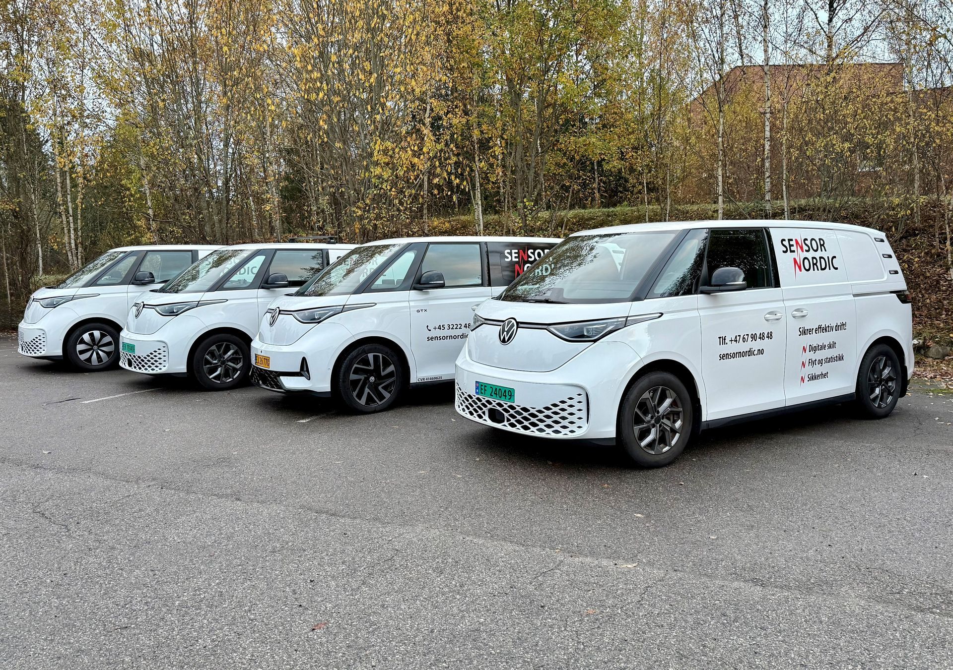 Fire hvite varebiler parkert på rad med grønne skilter. Varebilene står på en parkeringsplass med trær i bakgrunnen.
