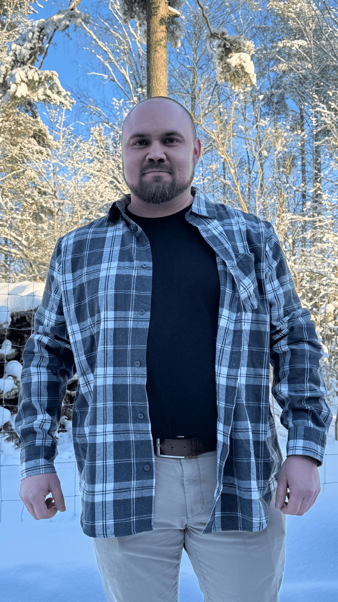Mann i rutete skjorte og svart t-skjorte står i en snødekt hage.