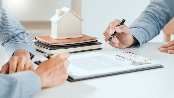 Due persone, sedute a una scrivania, esaminano il modellino di una casa e alcuni documenti, una delle quali scrive con una penna.