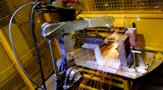 Welding robot at work, sparks flying, inside a yellow fenced enclosure.
