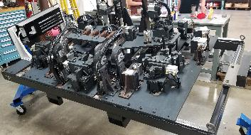 A complex, dark-colored industrial assembly on a metal base, supported by blue wheeled stands, in a workshop.