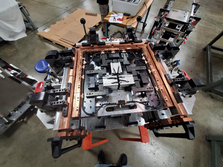 Industrial metalworking jig with copper-colored frame, surrounded by clamping mechanisms.