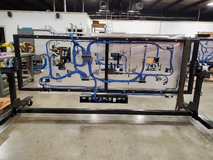 Black metal frame holding a wiring harness on a gray panel; blue wires, shop floor setting.