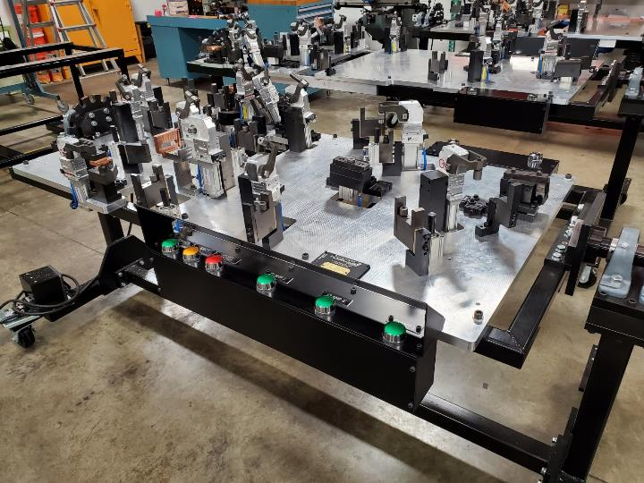 Metal fixture with clamps, buttons, and lights, likely for manufacturing, set on a rolling frame in a workshop.