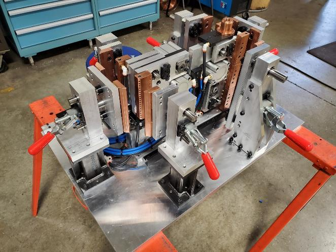 Industrial welding fixture, aluminum base, clamping mechanisms, copper contacts, red handles, blue wiring, workshop setting.