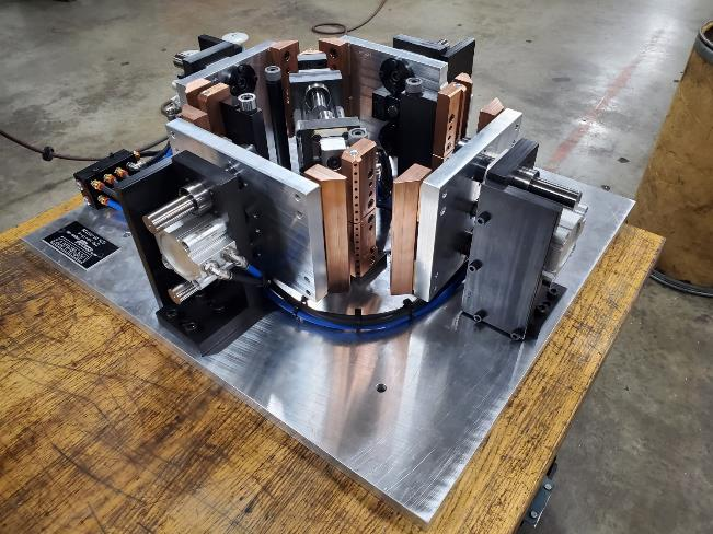 Machine with copper components, mounted on a square metal plate, resting on a wooden surface.