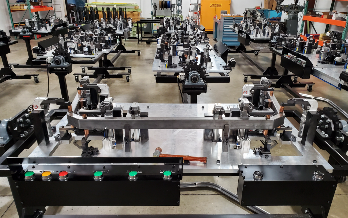 Workshop interior with multiple metal jigs on tables. A large assembly in the foreground has control buttons.