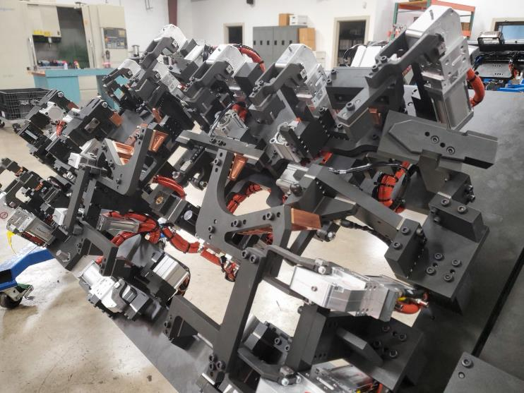 A complex industrial jig with multiple metal arms, clamps, and red wiring.