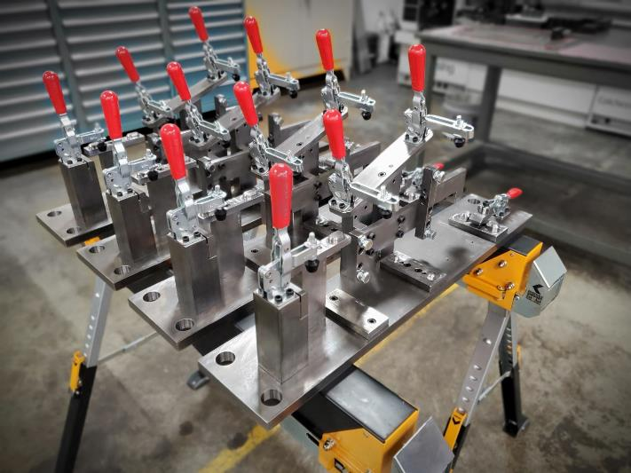 Metal jigs with red handles, mounted on a work table, used for holding parts in place during a manufacturing process.