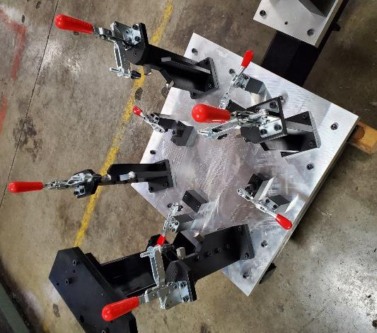 Metal jig with multiple toggle clamps, red handles, on a factory floor.