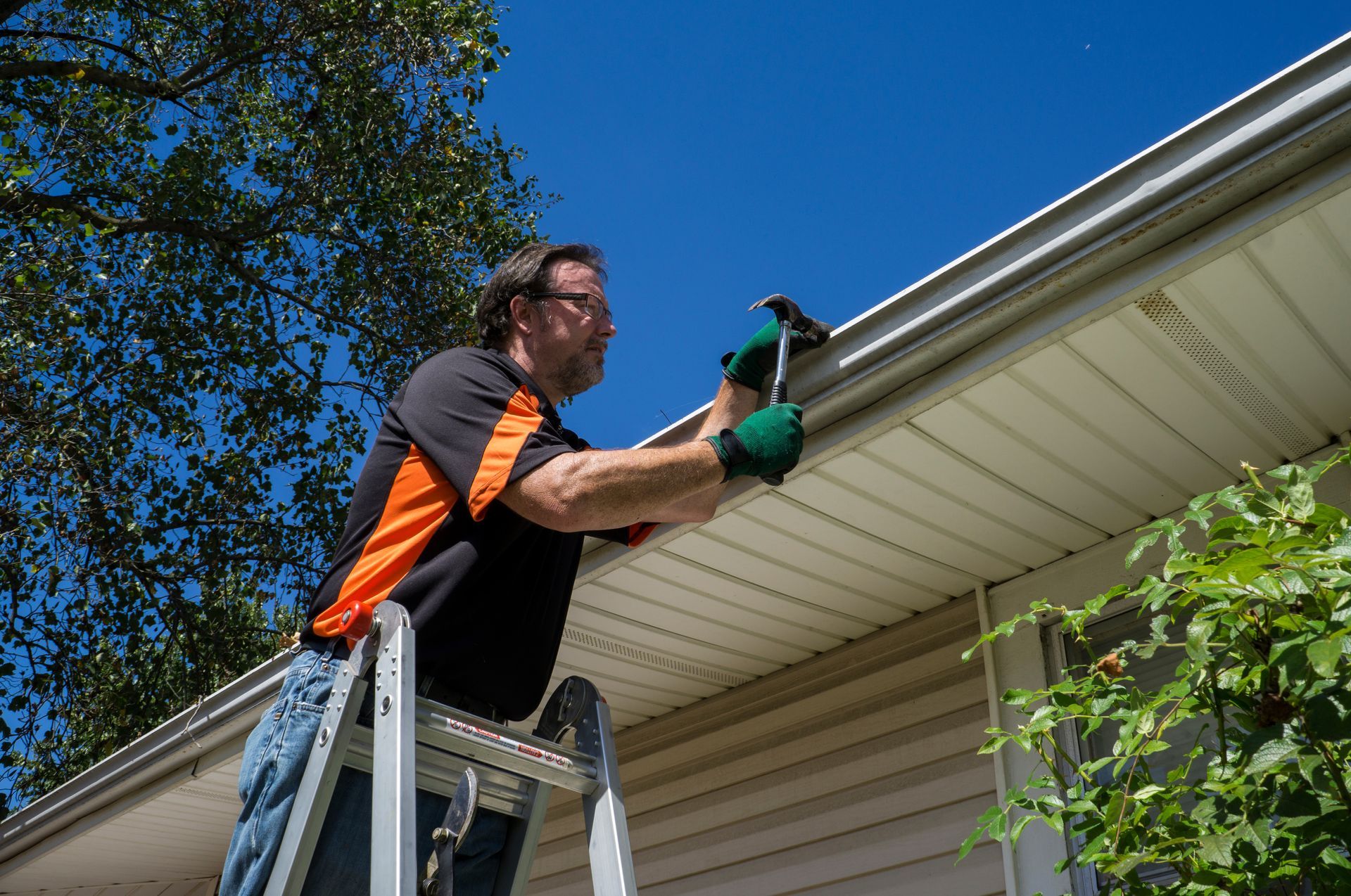 Person cleaning gutters with a green tool on a ladder beside a house roofline Person cleaning gutters with a green tool on a ladder beside a house roofline