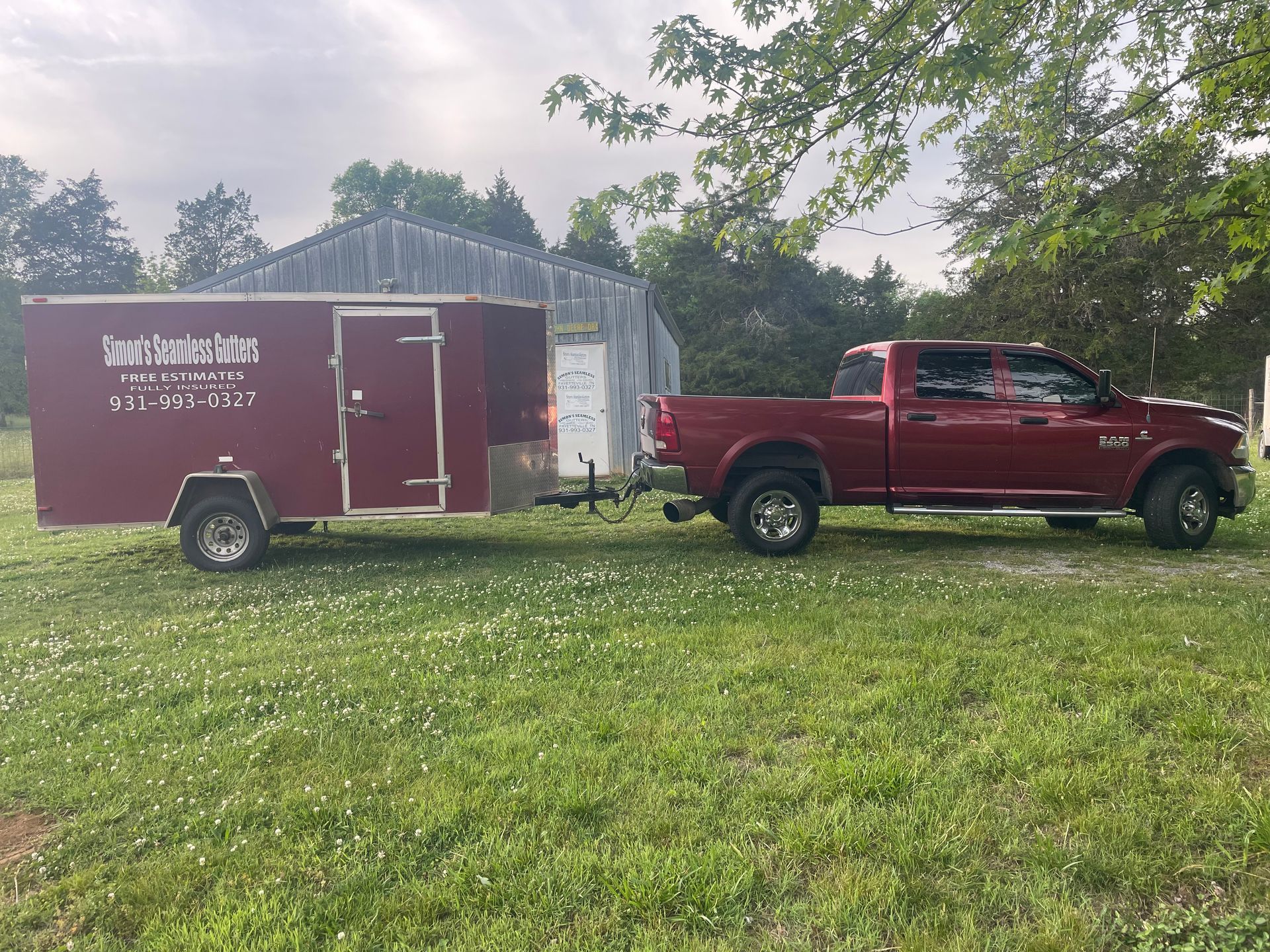 Red pickup truck hitched to a maroon enclosed trailer on a grassy field with trees in the background