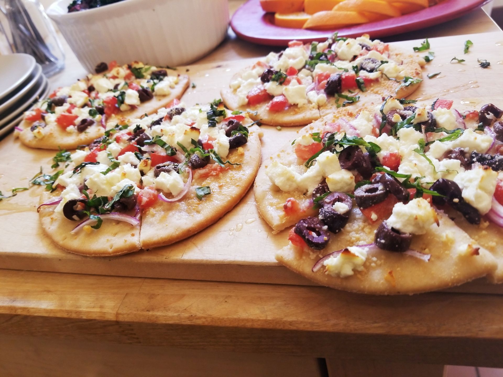 Mini pizzas topped with cheese, olives, herbs, and diced vegetables on a wooden cutting board