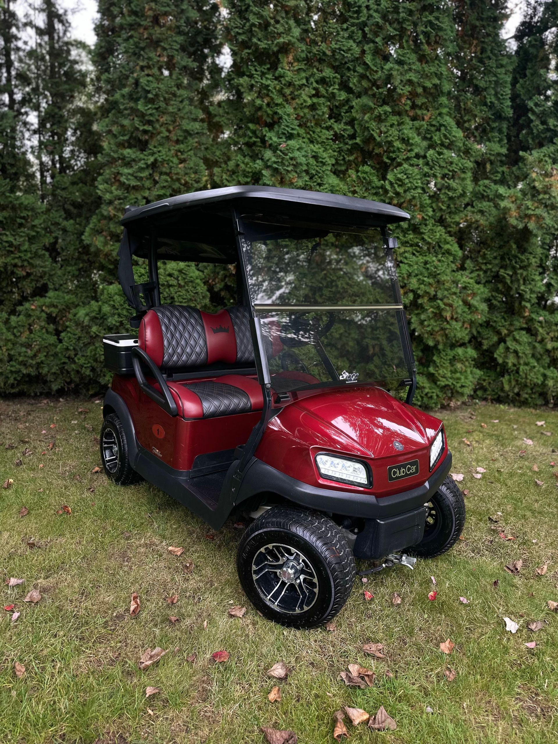 A red golf cart is parked in the grass in front of trees.