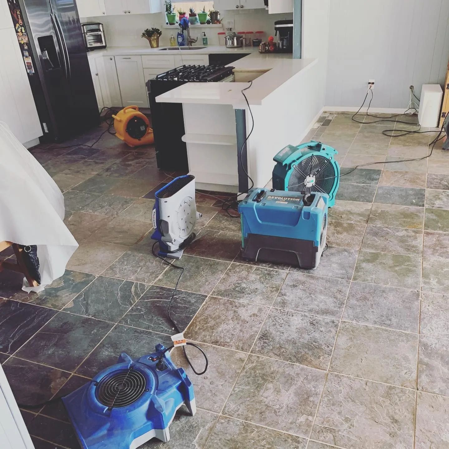 Flooded kitchen with dehumidifiers, fans, and cleaning equipment on a wet tile floor