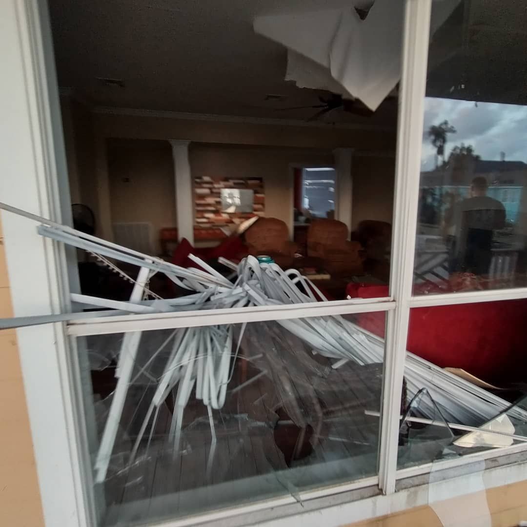 View through a broken window of a damaged home interior with furniture, debris, and reflection of a person.
