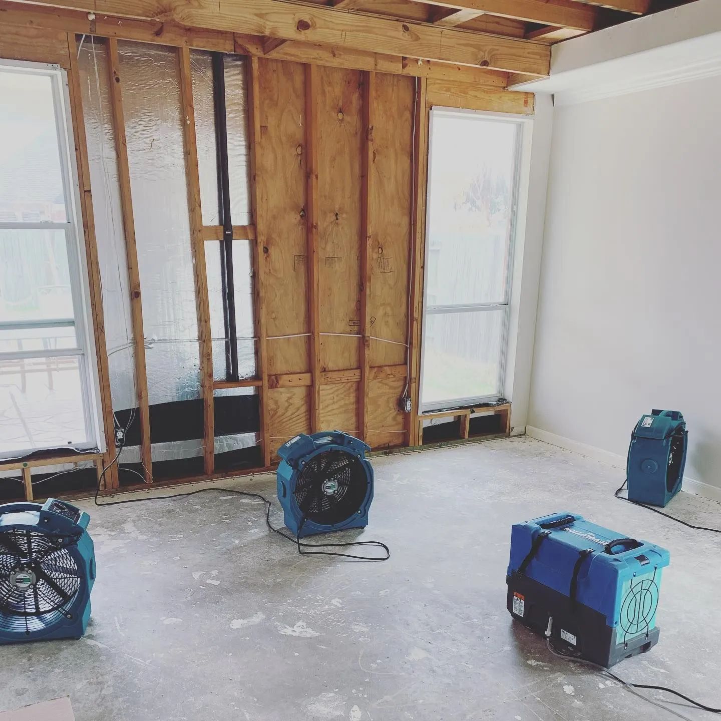 Partially renovated room with exposed framing, white walls, and three blue fans on the floor.