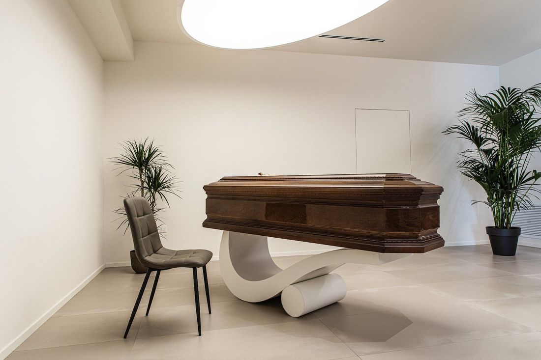 Un pianoforte a coda con base scultorea bianca in una stanza minimalista, affiancato da due piante in vaso e una sedia.