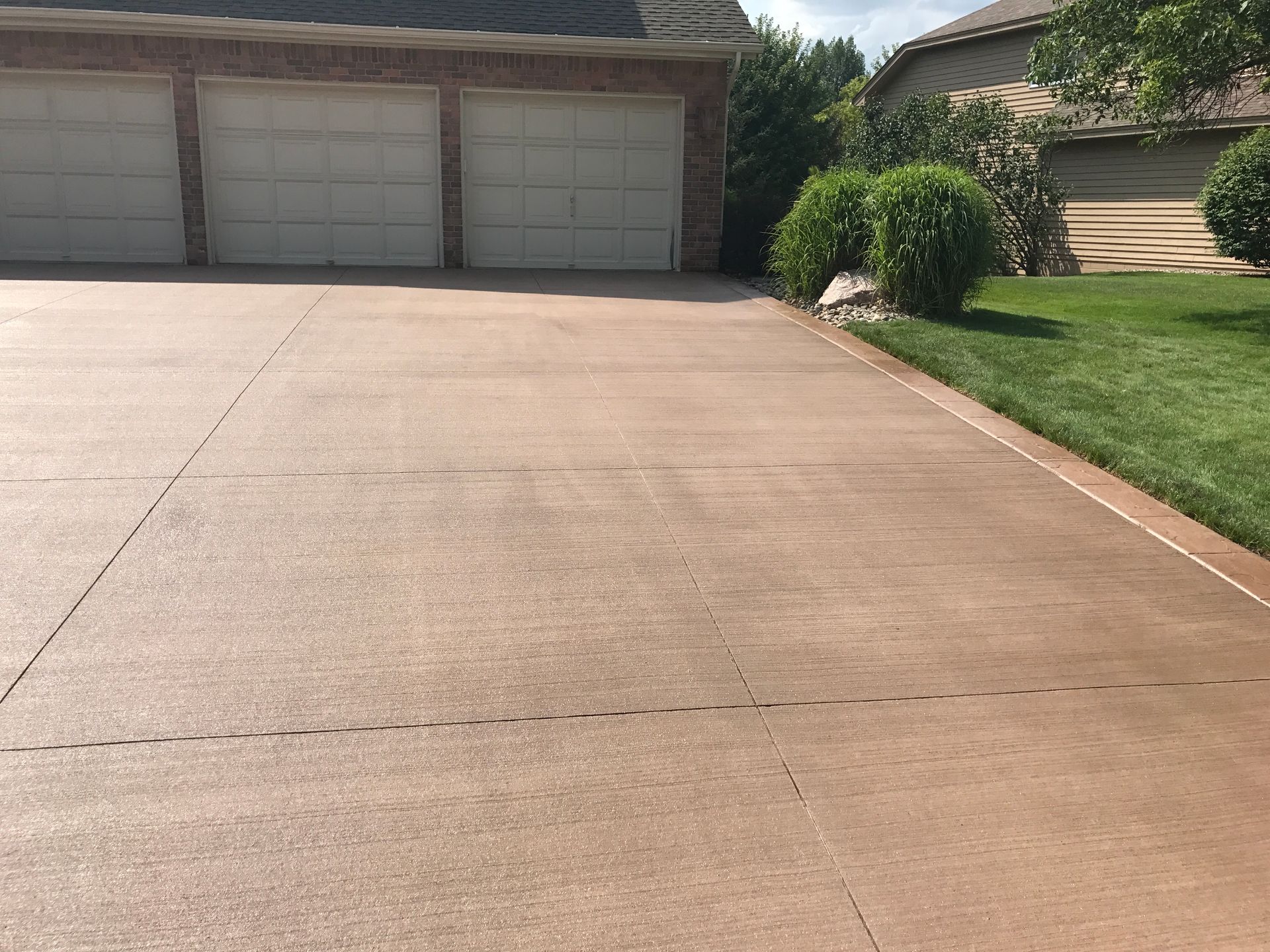Brown concrete driveway leading to a three-car garage. Green grass and bushes on the right.