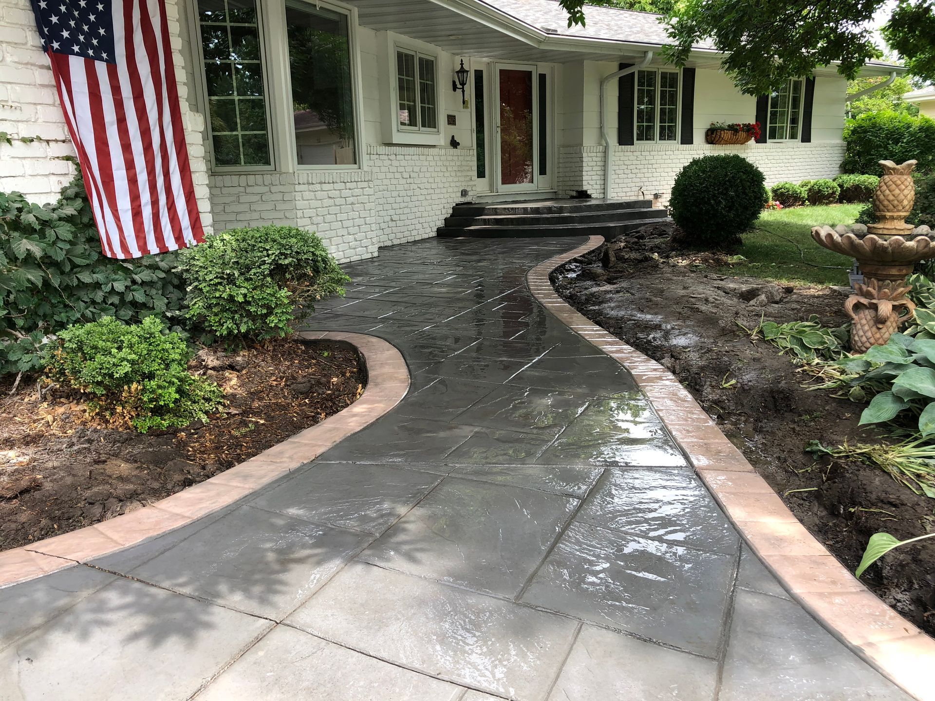 Brick walkway leading to a white brick house with an American flag and a fountain.