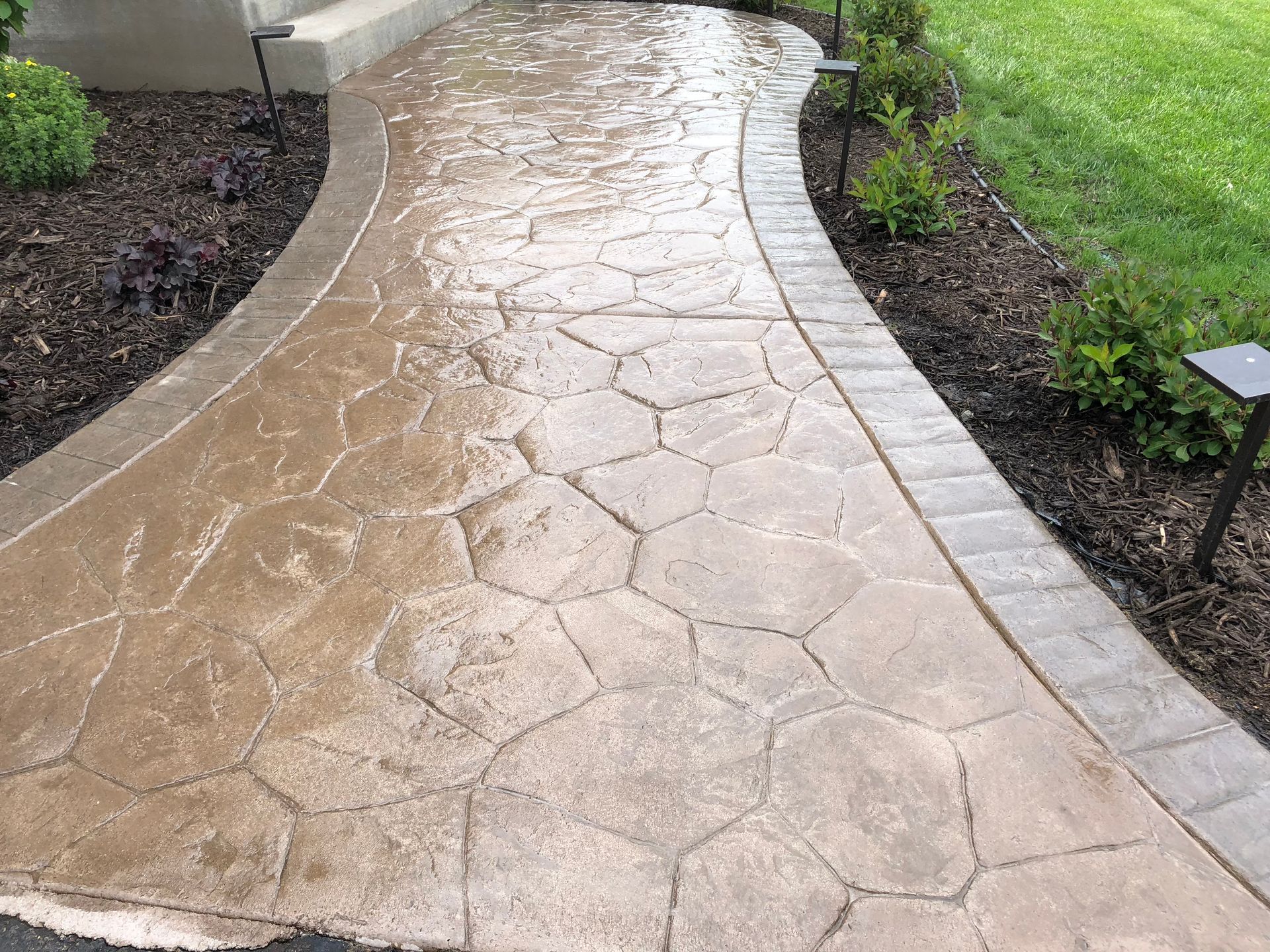 Stamped concrete walkway with stone-like pattern, bordered by brick and landscaping, leading to a house entrance.