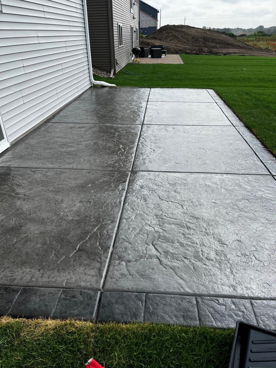 Concrete patio with textured surface, bordered by gray pavers and green grass, next to a white house.