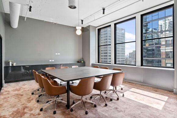 A modern conference room with a large square table, brown leather swivel chairs, and large windows overlooking a city.