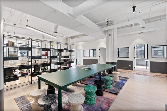 A modern office meeting space featuring a long green table, textured stools, and open shelving on a patterned rug.