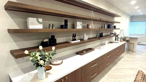 A modern Dermatology interior featuring minimalist wooden floating shelves displaying products above a long white counter.