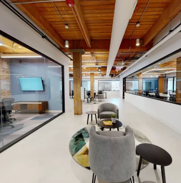Modern open-plan office lounge with grey armchairs, a wooden ceiling, and glass-enclosed meeting rooms.