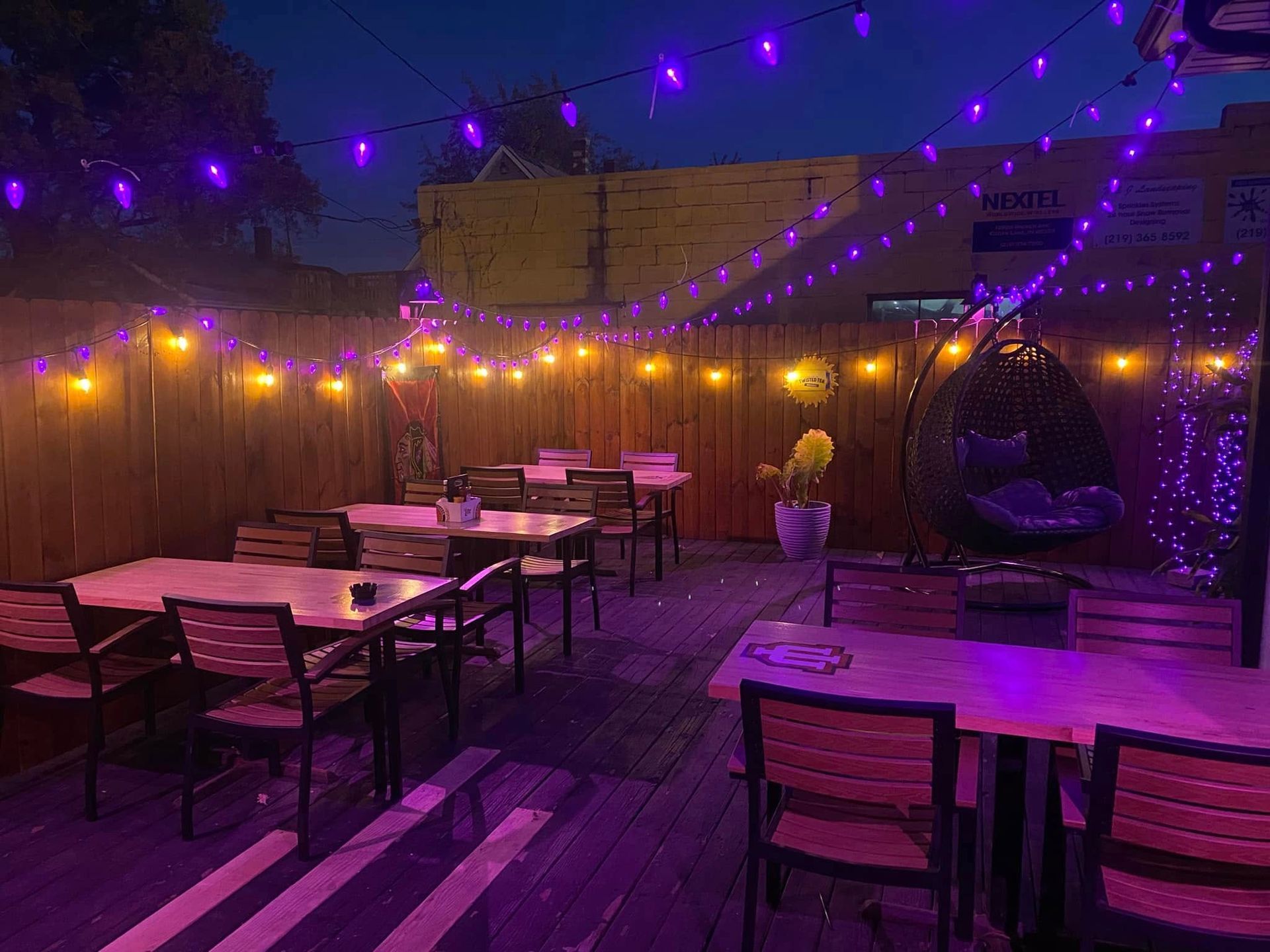 Outdoor patio at night, string lights overhead, wooden tables and chairs, wood fence.