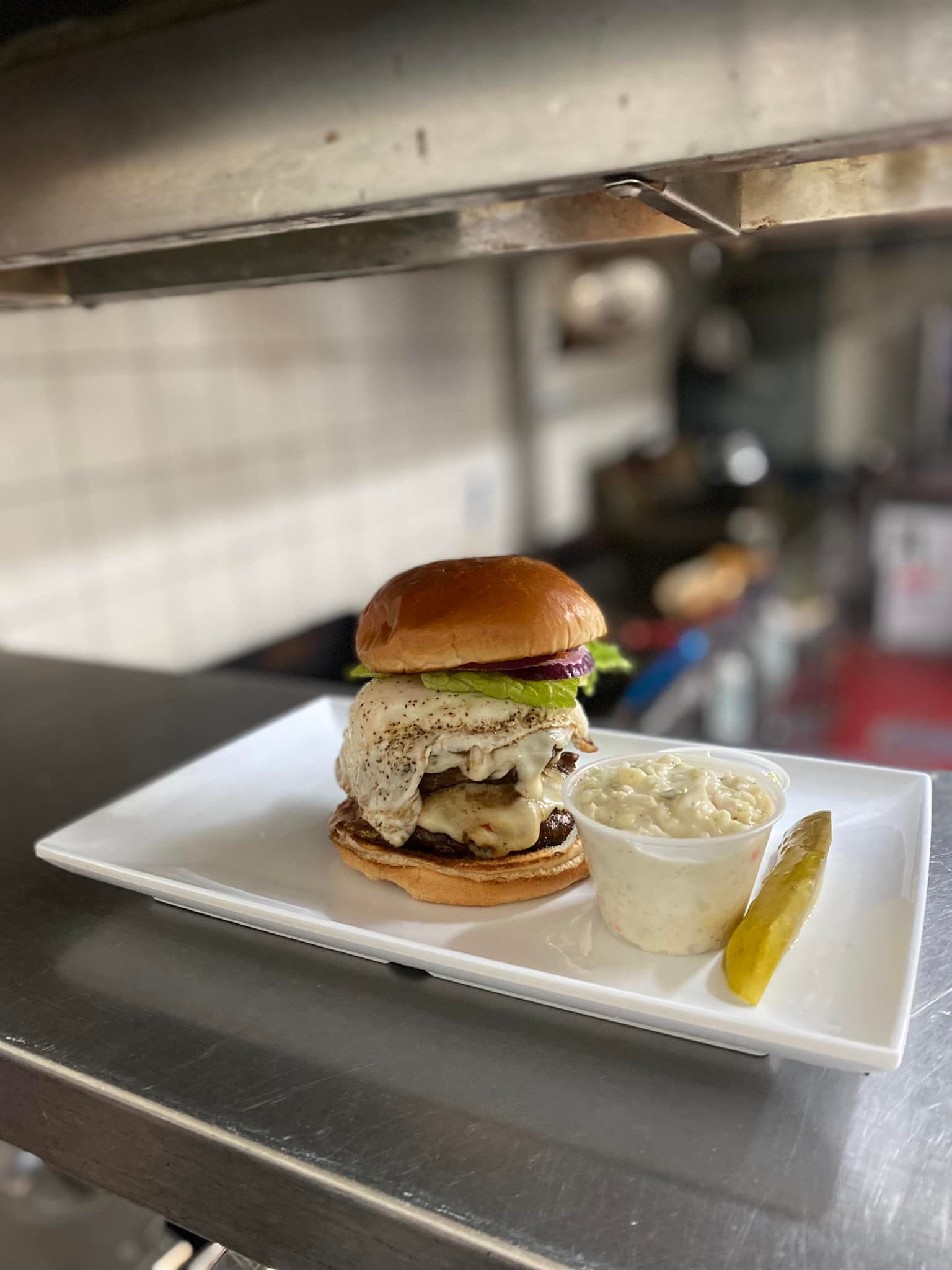 Double-stacked burger with cheese, lettuce, and bun, served with coleslaw and a pickle on a white rectangular plate.