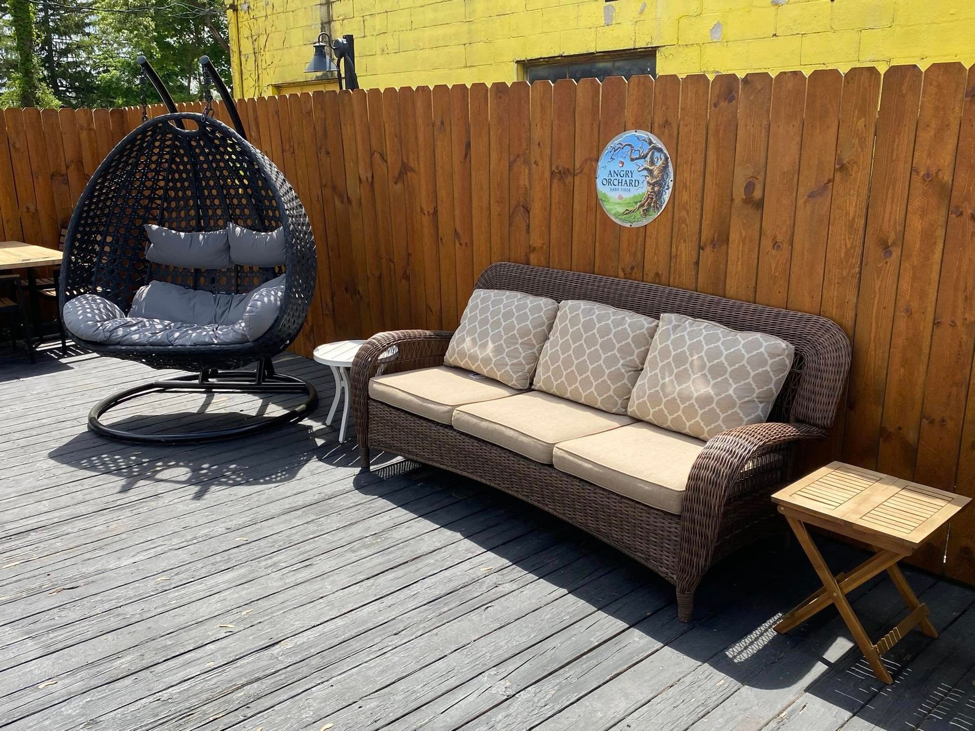 Outdoor patio with wicker sofa, hanging chair, wooden fence, and folding side table.
