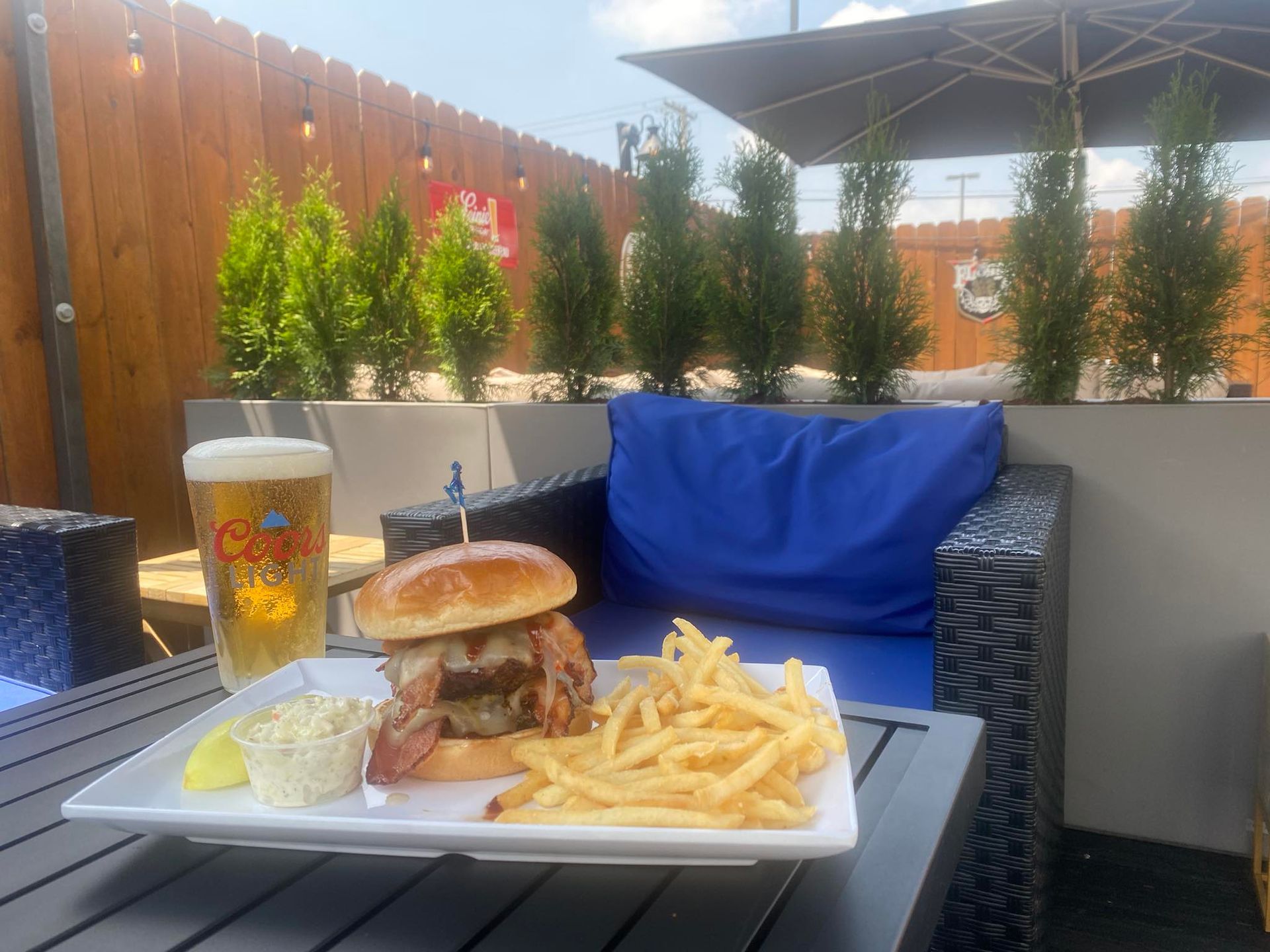 Burger, fries, and beer on an outdoor patio, blue cushions, wooden fence, sunny day.