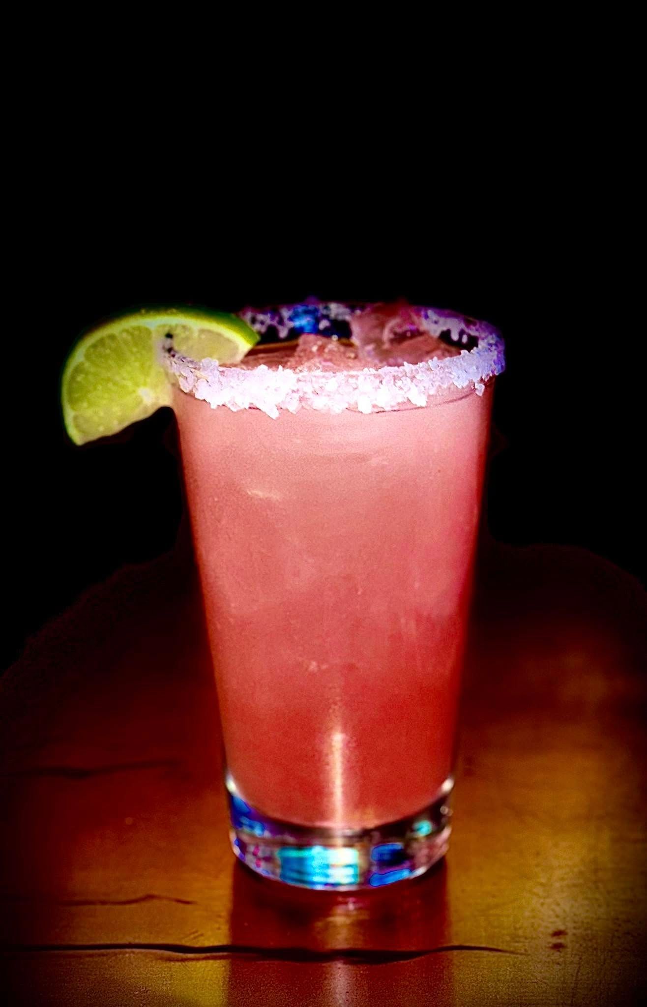 Pink cocktail in a salt-rimmed glass, garnished with lime wedge, on a table against a dark background.